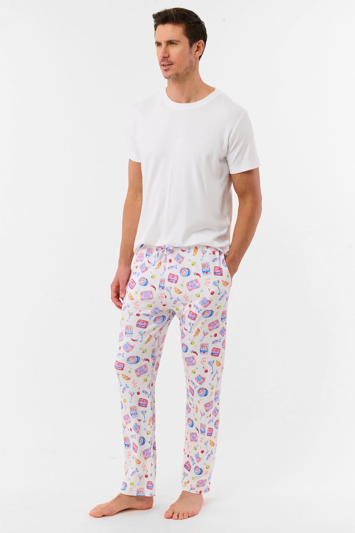 A man stands barefoot against a plain background, wearing the Roller Rabbit Mens Tinned or Twist Milo Set—a Pima cotton T-shirt paired with colorful drawstring waist pants featuring cartoon food patterns.