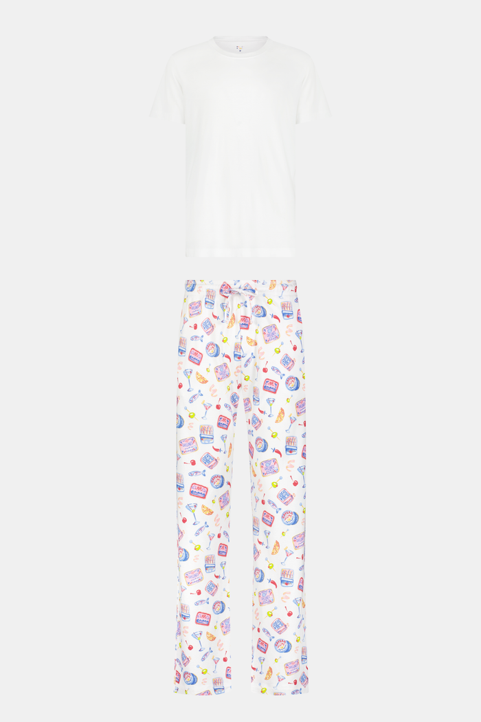 A man stands barefoot against a plain background, wearing the Roller Rabbit Mens Tinned or Twist Milo Set—a Pima cotton T-shirt paired with colorful drawstring waist pants featuring cartoon food patterns.