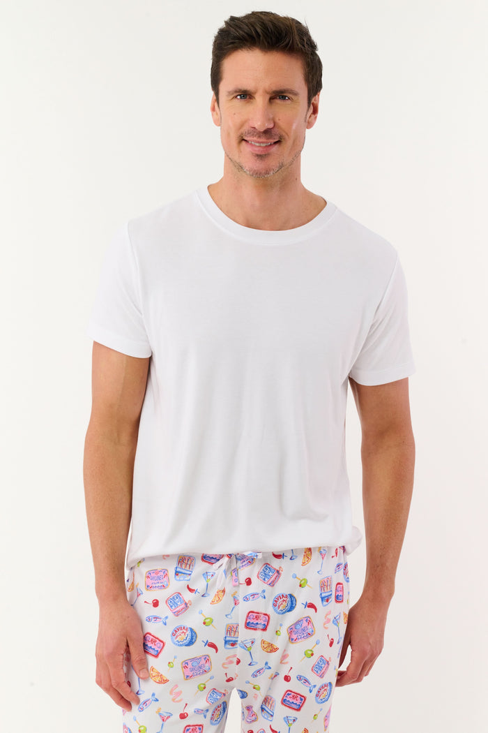 A man stands barefoot against a plain background, wearing the Roller Rabbit Mens Tinned or Twist Milo Set—a Pima cotton T-shirt paired with colorful drawstring waist pants featuring cartoon food patterns.