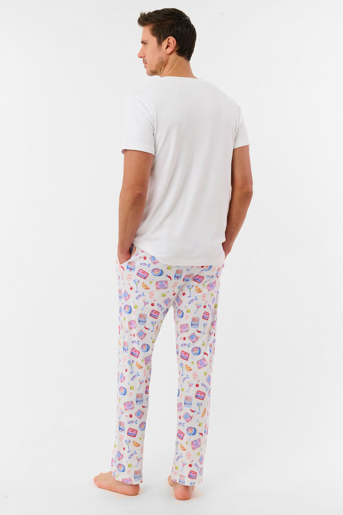 A man stands barefoot against a plain background, wearing the Roller Rabbit Mens Tinned or Twist Milo Set—a Pima cotton T-shirt paired with colorful drawstring waist pants featuring cartoon food patterns.