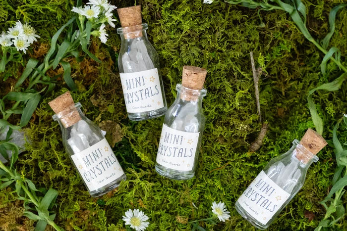 Four small clear glass bottles with cork stoppers and white labels reading 'MINI CRYSTALS' are arranged on green moss with small white flowers around them.