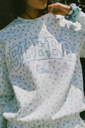 A person wearing a white Roller Rabbit Mini Monkey Blossom Andie Sweatshirt, featuring small blue and green Mini Monkey Blossom prints and large SLEEP Club lettering, stands outdoors, partially in sunlight. Their face is mostly out of frame.