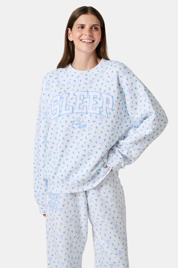 A person wearing a white Roller Rabbit Mini Monkey Blossom Andie Sweatshirt, featuring small blue and green Mini Monkey Blossom prints and large SLEEP Club lettering, stands outdoors, partially in sunlight. Their face is mostly out of frame.