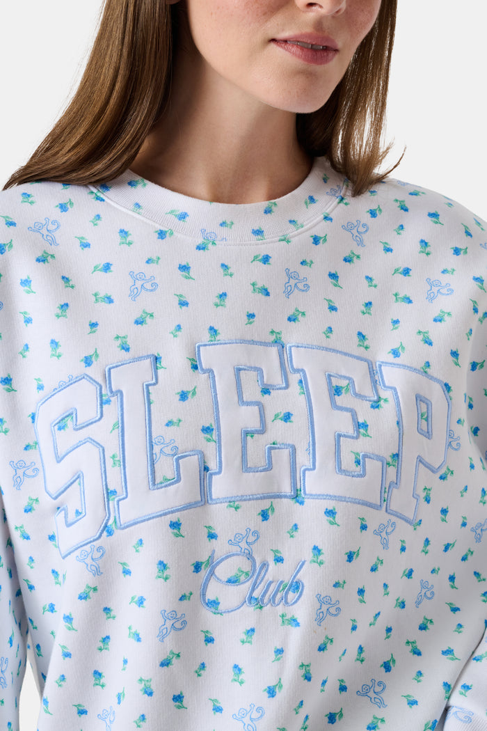 A person wearing a white Roller Rabbit Mini Monkey Blossom Andie Sweatshirt, featuring small blue and green Mini Monkey Blossom prints and large SLEEP Club lettering, stands outdoors, partially in sunlight. Their face is mostly out of frame.