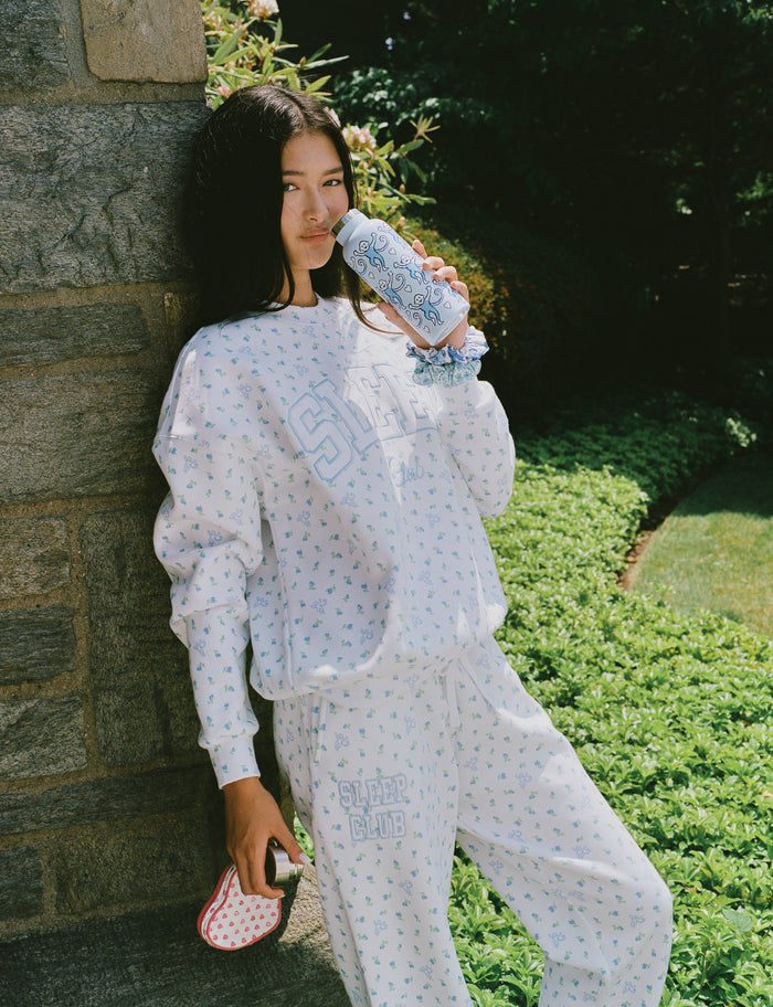 A person wearing a white Roller Rabbit Mini Monkey Blossom Andie Sweatshirt, featuring small blue and green Mini Monkey Blossom prints and large SLEEP Club lettering, stands outdoors, partially in sunlight. Their face is mostly out of frame.