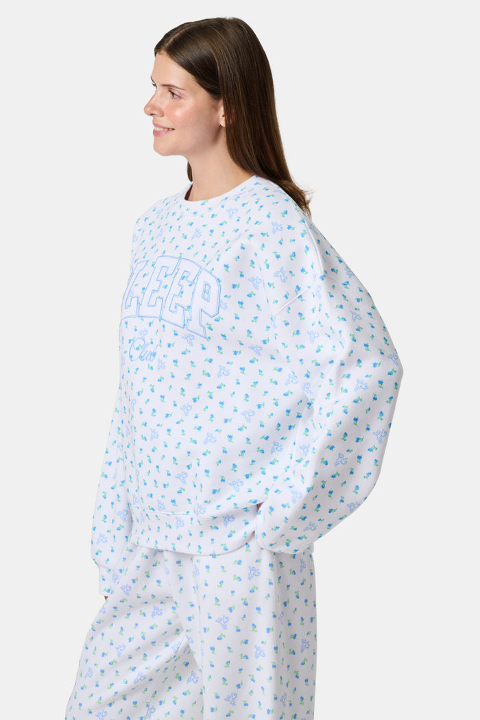 A person wearing a white Roller Rabbit Mini Monkey Blossom Andie Sweatshirt, featuring small blue and green Mini Monkey Blossom prints and large SLEEP Club lettering, stands outdoors, partially in sunlight. Their face is mostly out of frame.
