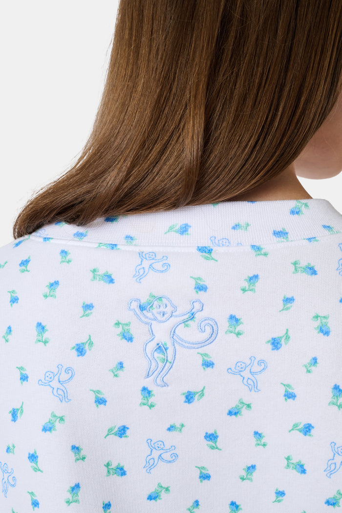 A person wearing a white Roller Rabbit Mini Monkey Blossom Andie Sweatshirt, featuring small blue and green Mini Monkey Blossom prints and large SLEEP Club lettering, stands outdoors, partially in sunlight. Their face is mostly out of frame.