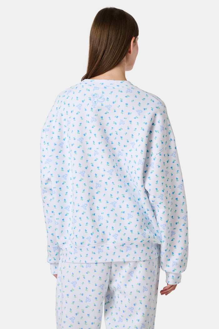 A person wearing a white Roller Rabbit Mini Monkey Blossom Andie Sweatshirt, featuring small blue and green Mini Monkey Blossom prints and large SLEEP Club lettering, stands outdoors, partially in sunlight. Their face is mostly out of frame.