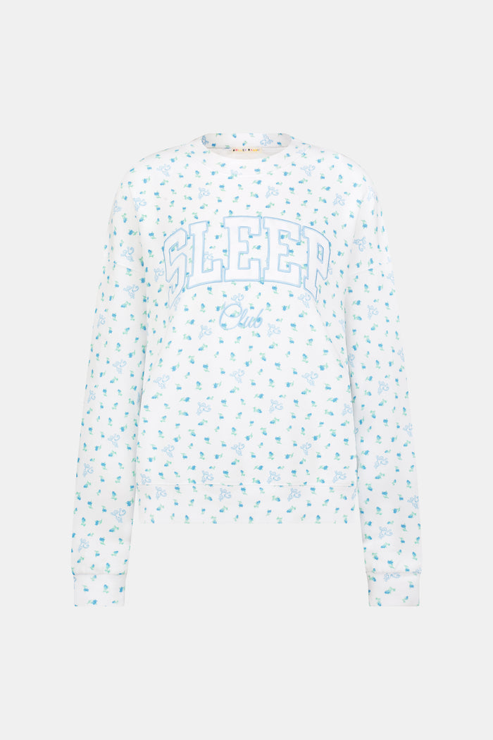A person wearing a white Roller Rabbit Mini Monkey Blossom Andie Sweatshirt, featuring small blue and green Mini Monkey Blossom prints and large SLEEP Club lettering, stands outdoors, partially in sunlight. Their face is mostly out of frame.