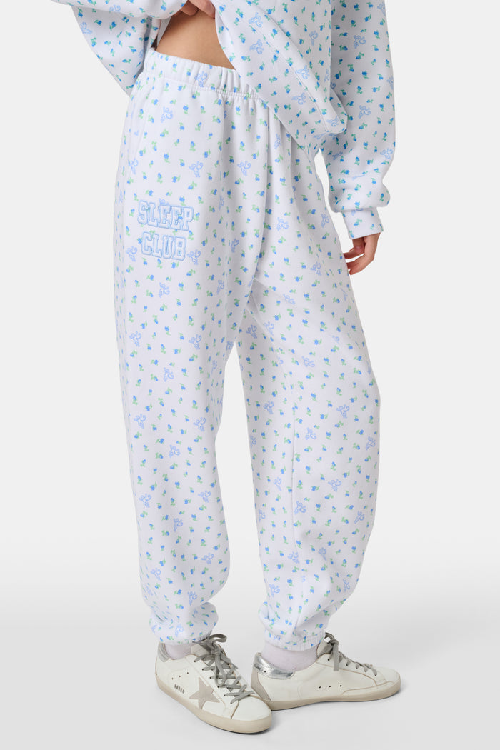 A person wearing a white Roller Rabbit Mini Monkey Blossom Andie Sweatshirt, featuring small blue and green Mini Monkey Blossom prints and large SLEEP Club lettering, stands outdoors, partially in sunlight. Their face is mostly out of frame.