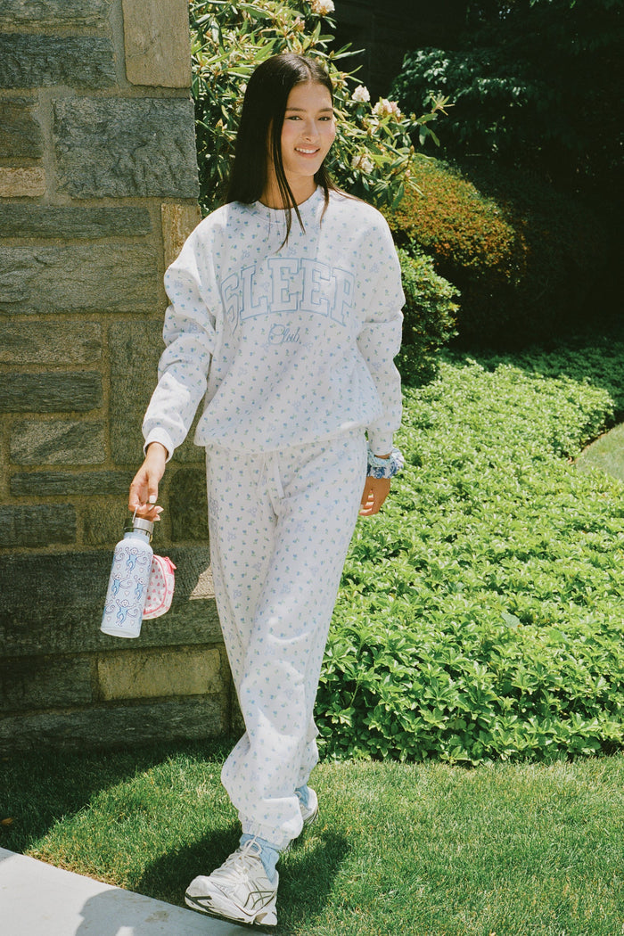 A person wearing a white Roller Rabbit Mini Monkey Blossom Andie Sweatshirt, featuring small blue and green Mini Monkey Blossom prints and large SLEEP Club lettering, stands outdoors, partially in sunlight. Their face is mostly out of frame.
