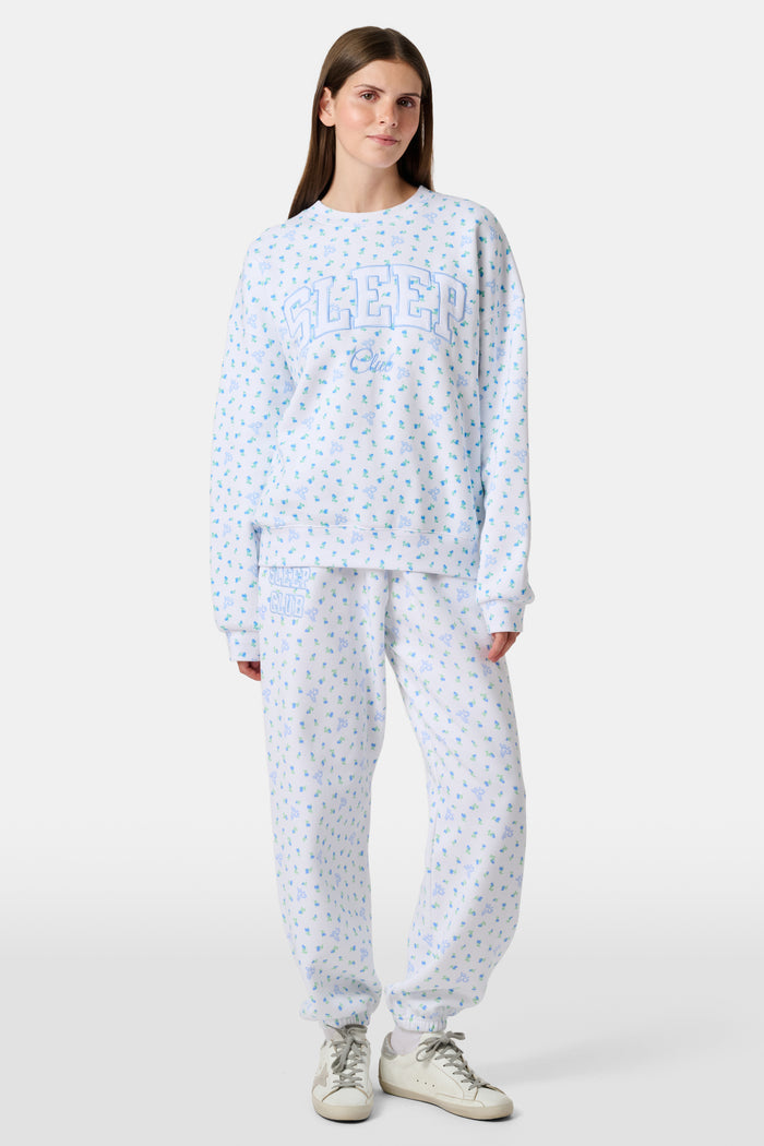 A person wearing a white Roller Rabbit Mini Monkey Blossom Andie Sweatshirt, featuring small blue and green Mini Monkey Blossom prints and large SLEEP Club lettering, stands outdoors, partially in sunlight. Their face is mostly out of frame.