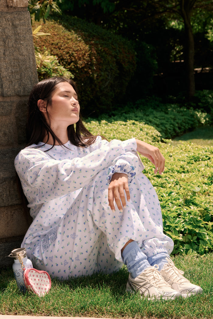 A person wearing a white Roller Rabbit Mini Monkey Blossom Andie Sweatshirt, featuring small blue and green Mini Monkey Blossom prints and large SLEEP Club lettering, stands outdoors, partially in sunlight. Their face is mostly out of frame.