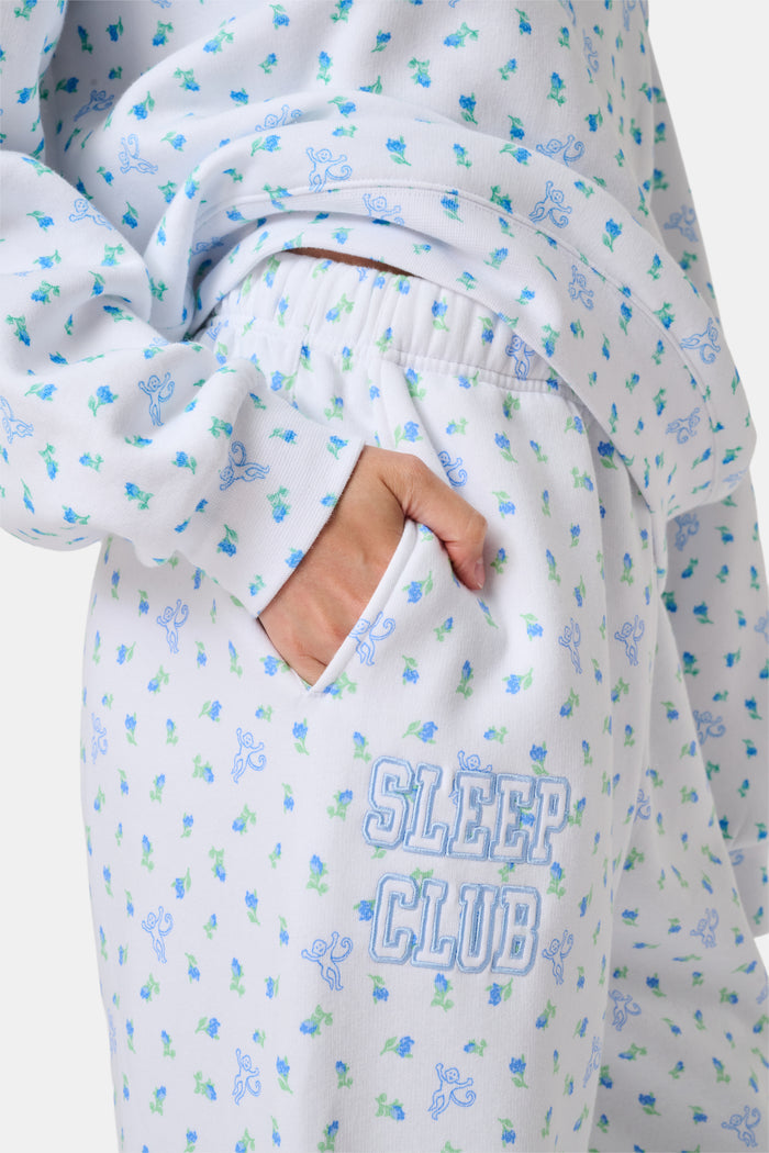 A person wearing a white Roller Rabbit Mini Monkey Blossom Andie Sweatshirt, featuring small blue and green Mini Monkey Blossom prints and large SLEEP Club lettering, stands outdoors, partially in sunlight. Their face is mostly out of frame.