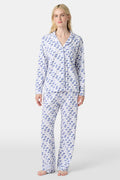 A woman stands barefoot in the Roller Rabbit Moby Long Sleeve Polo Pajamas, featuring a blue geometric-patterned white top and pants. Made from soft pima cotton, the comfy set is shown against a plain white background.