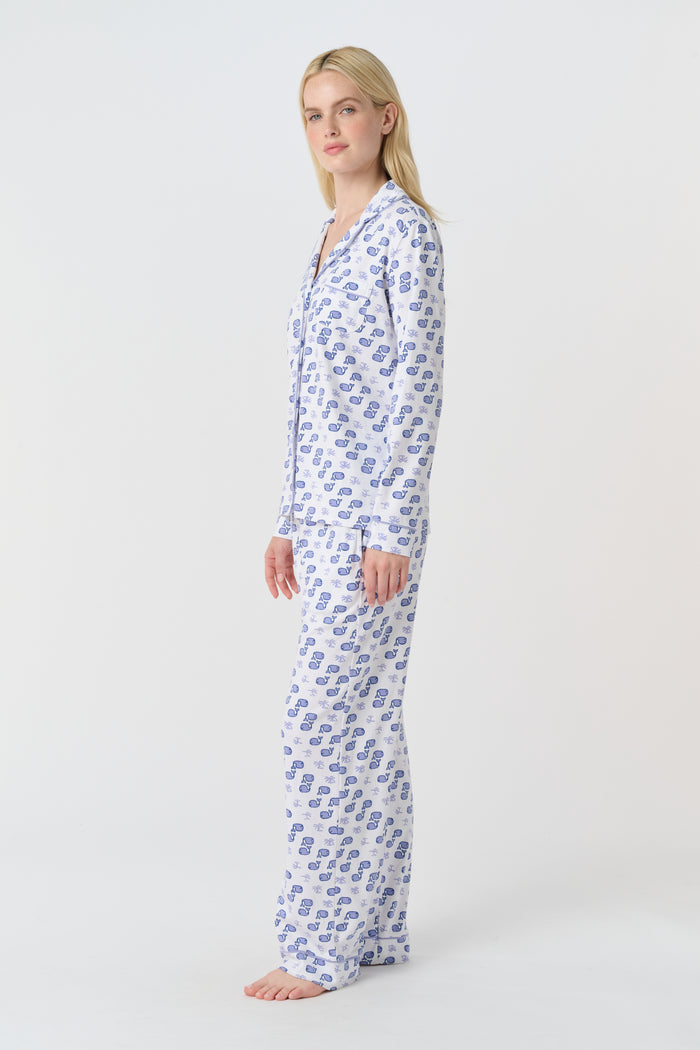A woman stands barefoot in the Roller Rabbit Moby Long Sleeve Polo Pajamas, featuring a blue geometric-patterned white top and pants. Made from soft pima cotton, the comfy set is shown against a plain white background.