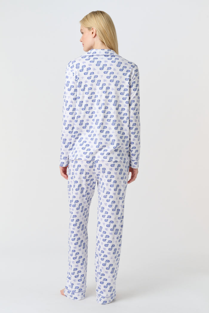 A woman stands barefoot in the Roller Rabbit Moby Long Sleeve Polo Pajamas, featuring a blue geometric-patterned white top and pants. Made from soft pima cotton, the comfy set is shown against a plain white background.