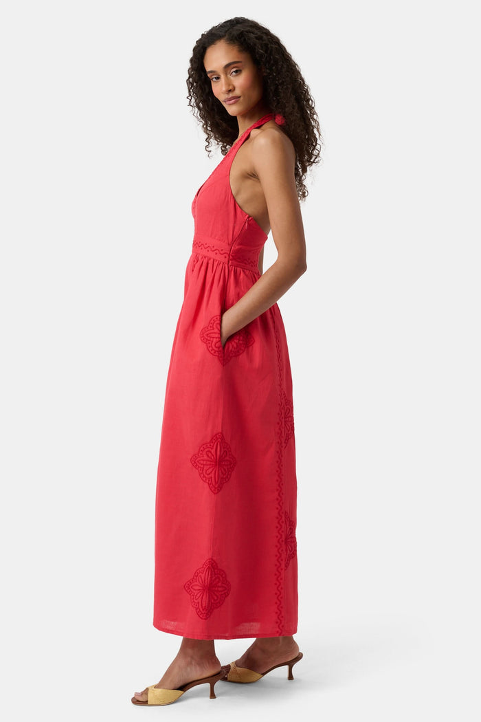 A woman with long curly hair wears the Roller Rabbit Monaco Geo Penelope Dress, a sleeveless, halter-neck, ankle-length red dress with subtle embroidery. She stands forward in open-toed, low-heeled sandals against a plain white background.