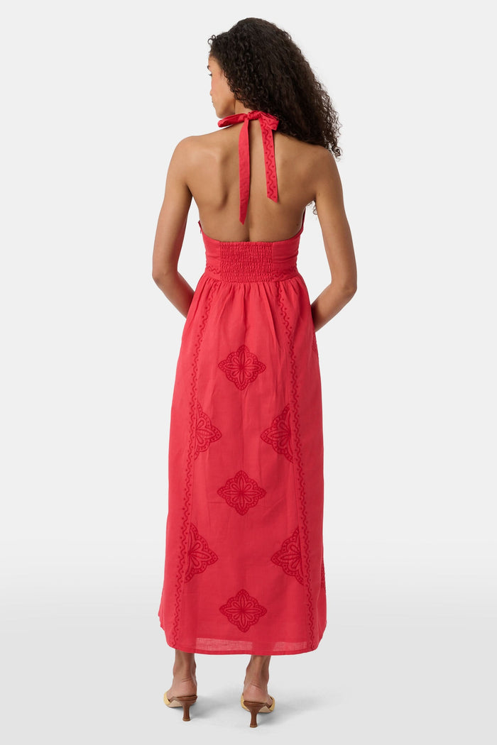 A woman with long curly hair wears the Roller Rabbit Monaco Geo Penelope Dress, a sleeveless, halter-neck, ankle-length red dress with subtle embroidery. She stands forward in open-toed, low-heeled sandals against a plain white background.