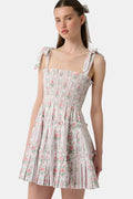 A young woman wears the Roller Rabbit Monkey Block Stripe Ribbon Charlotte Nap Dress, featuring a smocked bodice, ruffled hem, and tied shoulder straps. Her straight hair is partly tied back with a matching ribbon against a plain white background.
