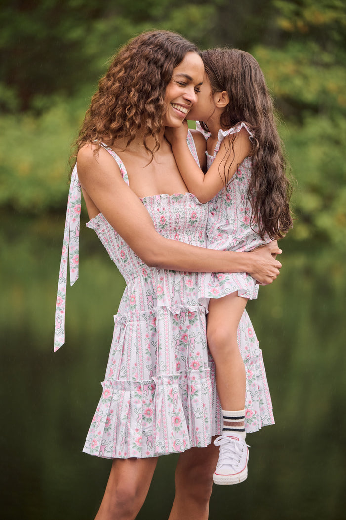 A young woman wears the Roller Rabbit Monkey Block Stripe Ribbon Charlotte Nap Dress, featuring a smocked bodice, ruffled hem, and tied shoulder straps. Her straight hair is partly tied back with a matching ribbon against a plain white background.
