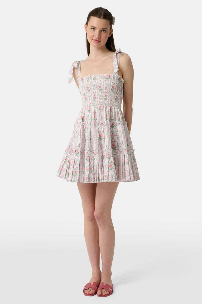 A young woman wears the Roller Rabbit Monkey Block Stripe Ribbon Charlotte Nap Dress, featuring a smocked bodice, ruffled hem, and tied shoulder straps. Her straight hair is partly tied back with a matching ribbon against a plain white background.