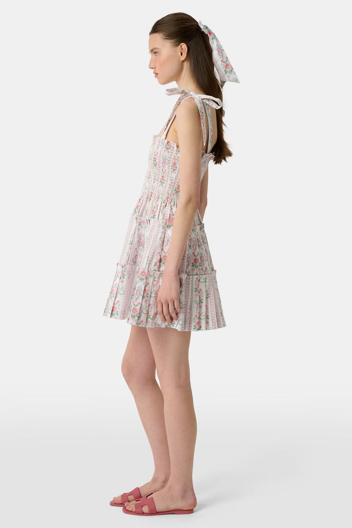 A young woman wears the Roller Rabbit Monkey Block Stripe Ribbon Charlotte Nap Dress, featuring a smocked bodice, ruffled hem, and tied shoulder straps. Her straight hair is partly tied back with a matching ribbon against a plain white background.
