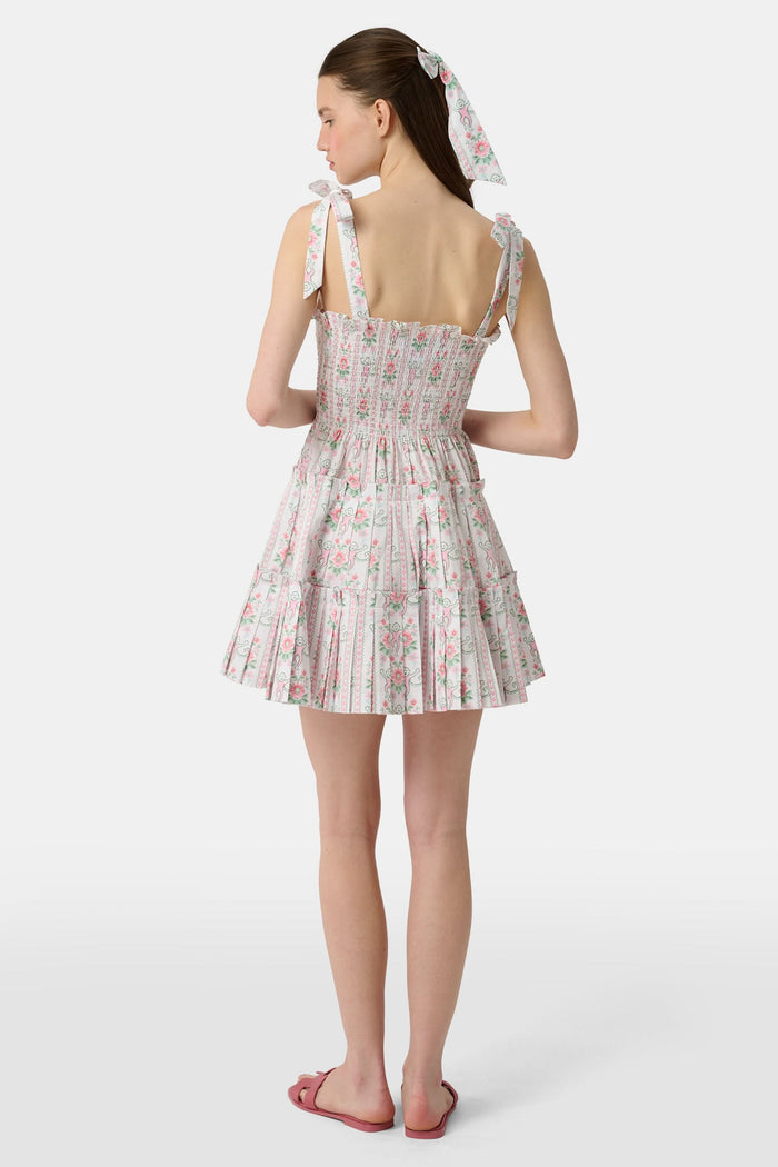 A young woman wears the Roller Rabbit Monkey Block Stripe Ribbon Charlotte Nap Dress, featuring a smocked bodice, ruffled hem, and tied shoulder straps. Her straight hair is partly tied back with a matching ribbon against a plain white background.