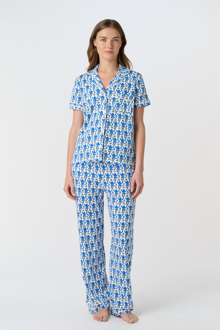 A woman stands barefoot against a plain white background wearing the Roller Rabbit Monkey Fiora Pajamas, a blue-and-white patterned short-sleeve polo pajama set with matching pants.