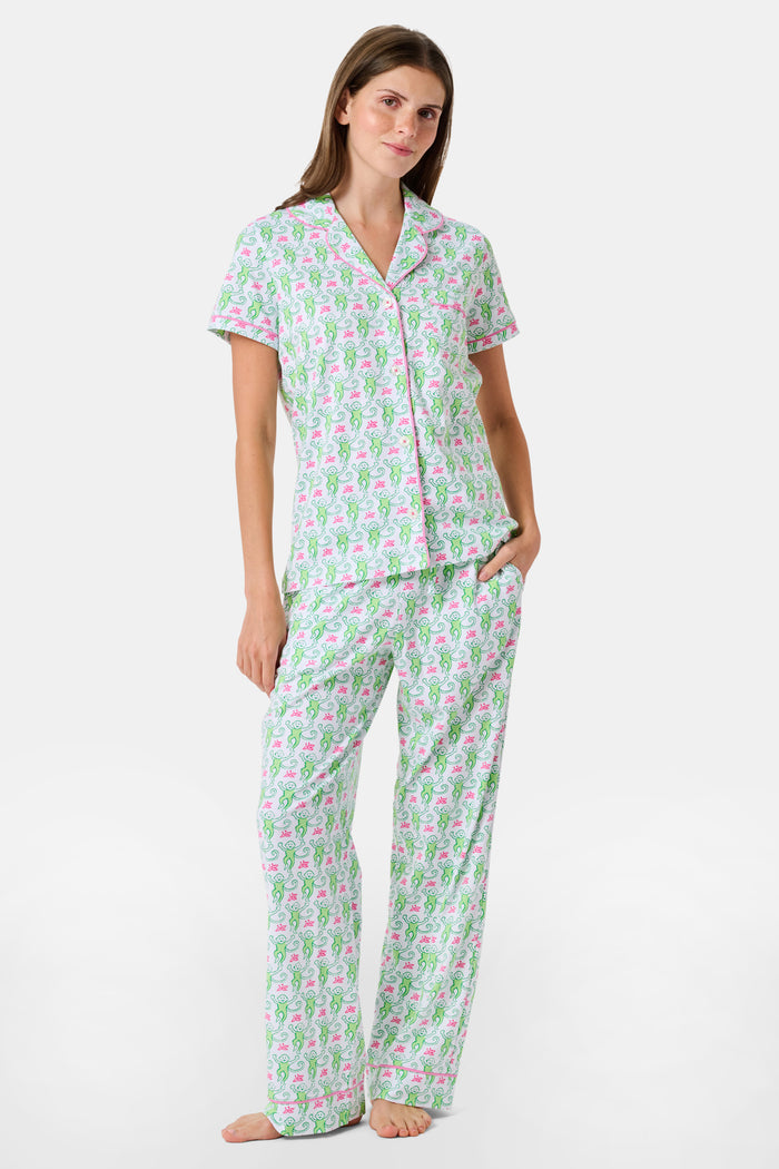 A woman stands barefoot against a plain white background, wearing the Roller Rabbit Monkey Fiora Pajamas—a matching set crafted from soft Pima cotton for extra comfort. The pajama set features a short-sleeve button-up top and long pants adorned with a pastel floral motif.
