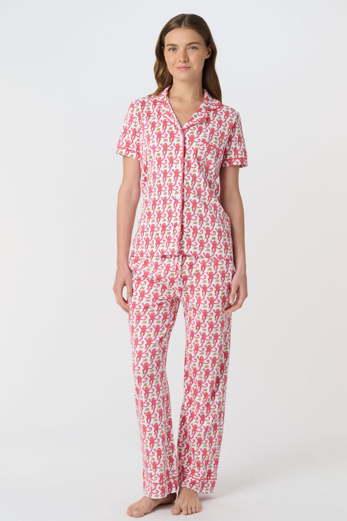 A woman stands barefoot against a plain white background, wearing the Roller Rabbit Monkey Fiora Pajamas set, which features a short-sleeve, button-up top and long pants.