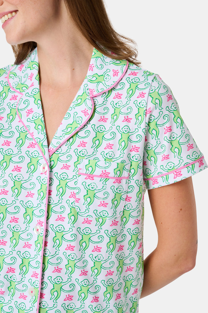 A woman stands barefoot against a plain white background, wearing the Roller Rabbit Monkey Fiora Pajamas—a matching set crafted from soft Pima cotton for extra comfort. The pajama set features a short-sleeve button-up top and long pants adorned with a pastel floral motif.