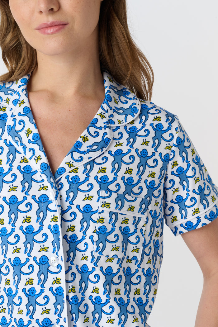 A woman stands barefoot against a plain white background wearing the Roller Rabbit Monkey Fiora Pajamas, a blue-and-white patterned short-sleeve polo pajama set with matching pants.