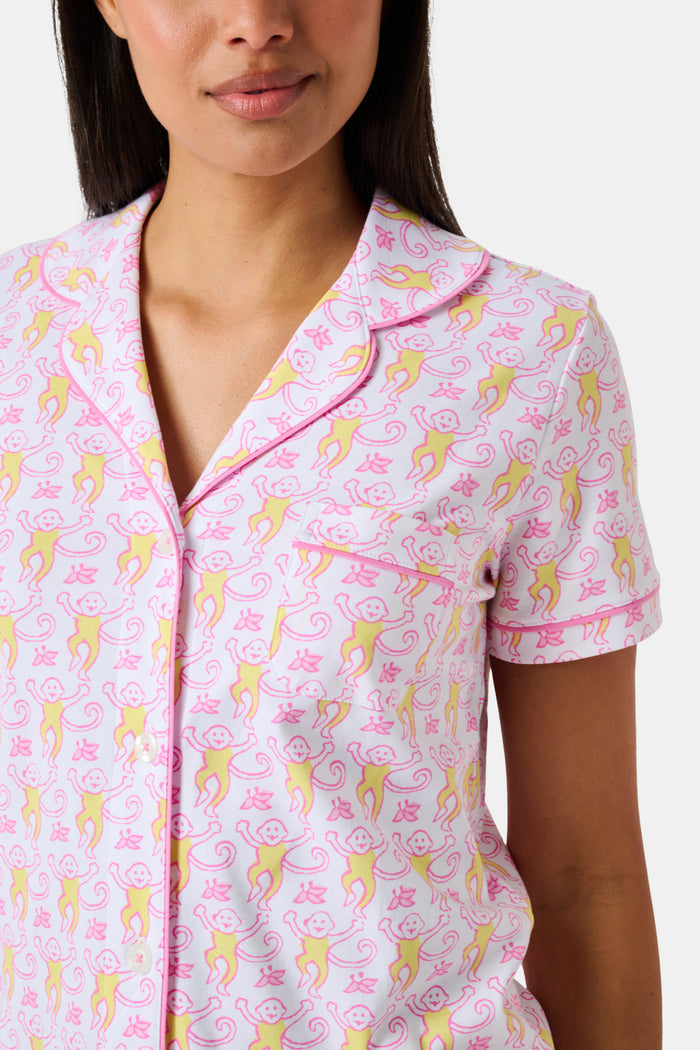 Introducing the Monkey Fiora Pajamas from Roller Rabbit—crafted from fabric adorned with a delightful pattern of pink and yellow monkeys with curled tails and smiling faces, interspersed with tiny leaves on a crisp white background. Perfect for adding playful charm to your sleepwear collection.