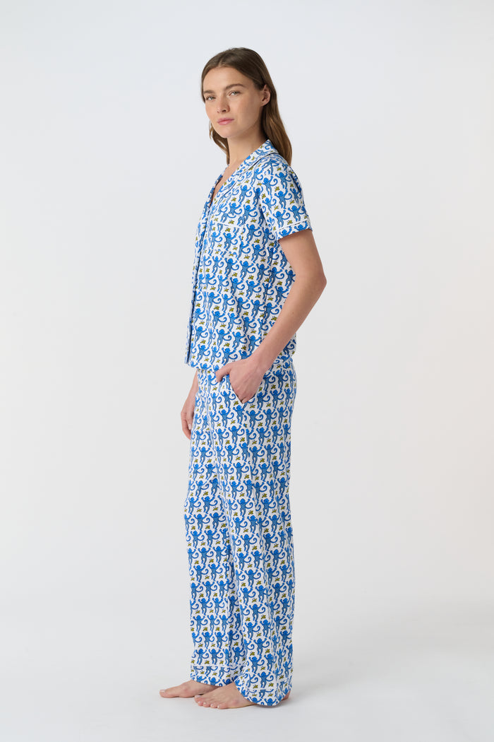 A woman stands barefoot against a plain white background wearing the Roller Rabbit Monkey Fiora Pajamas, a blue-and-white patterned short-sleeve polo pajama set with matching pants.