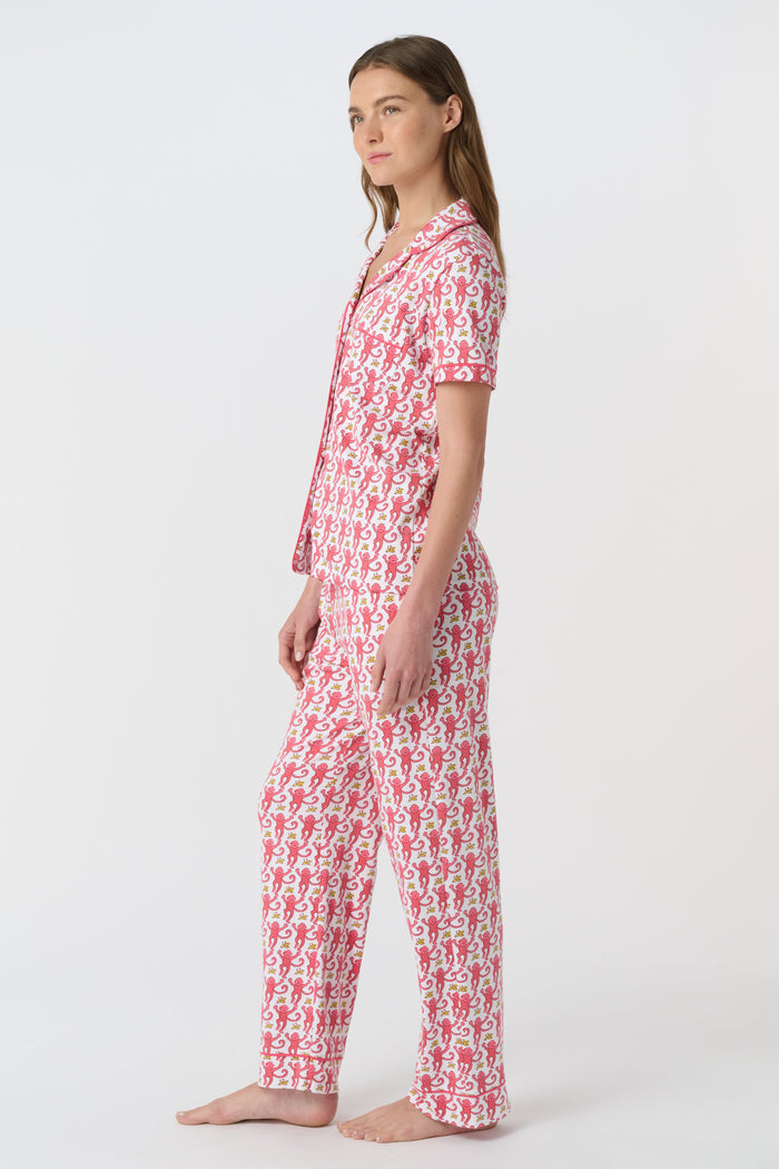 A woman stands barefoot against a plain white background, wearing the Roller Rabbit Monkey Fiora Pajamas set, which features a short-sleeve, button-up top and long pants.