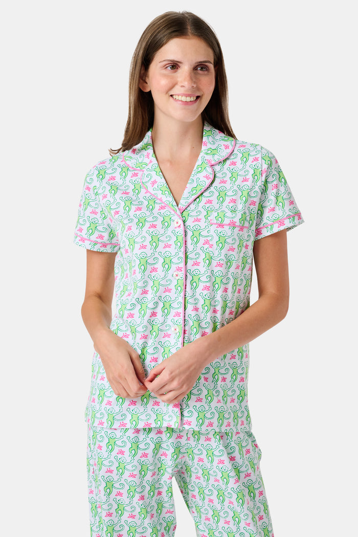 A woman stands barefoot against a plain white background, wearing the Roller Rabbit Monkey Fiora Pajamas—a matching set crafted from soft Pima cotton for extra comfort. The pajama set features a short-sleeve button-up top and long pants adorned with a pastel floral motif.