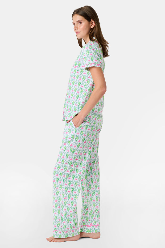 A woman stands barefoot against a plain white background, wearing the Roller Rabbit Monkey Fiora Pajamas—a matching set crafted from soft Pima cotton for extra comfort. The pajama set features a short-sleeve button-up top and long pants adorned with a pastel floral motif.