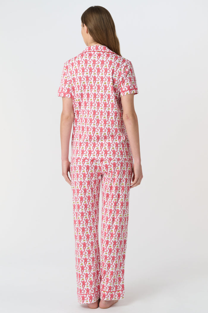 A woman stands barefoot against a plain white background, wearing the Roller Rabbit Monkey Fiora Pajamas set, which features a short-sleeve, button-up top and long pants.