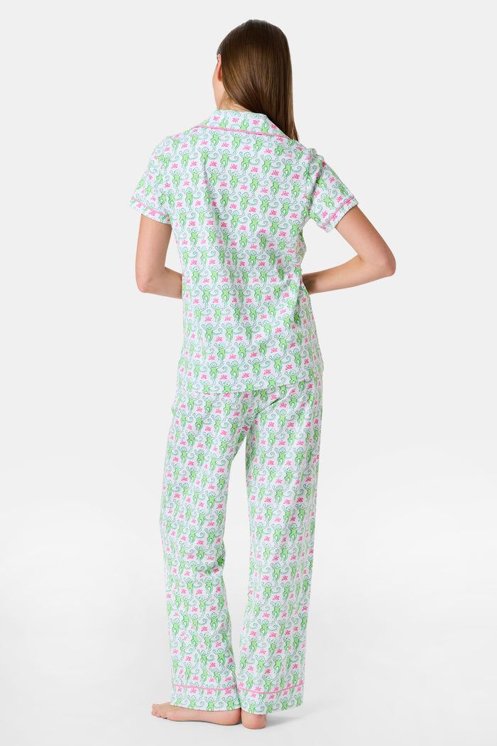 A woman stands barefoot against a plain white background, wearing the Roller Rabbit Monkey Fiora Pajamas—a matching set crafted from soft Pima cotton for extra comfort. The pajama set features a short-sleeve button-up top and long pants adorned with a pastel floral motif.