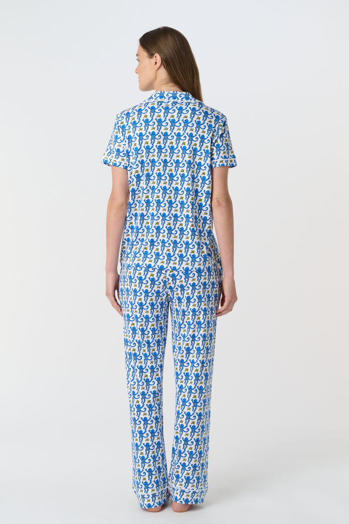 A woman stands barefoot against a plain white background wearing the Roller Rabbit Monkey Fiora Pajamas, a blue-and-white patterned short-sleeve polo pajama set with matching pants.