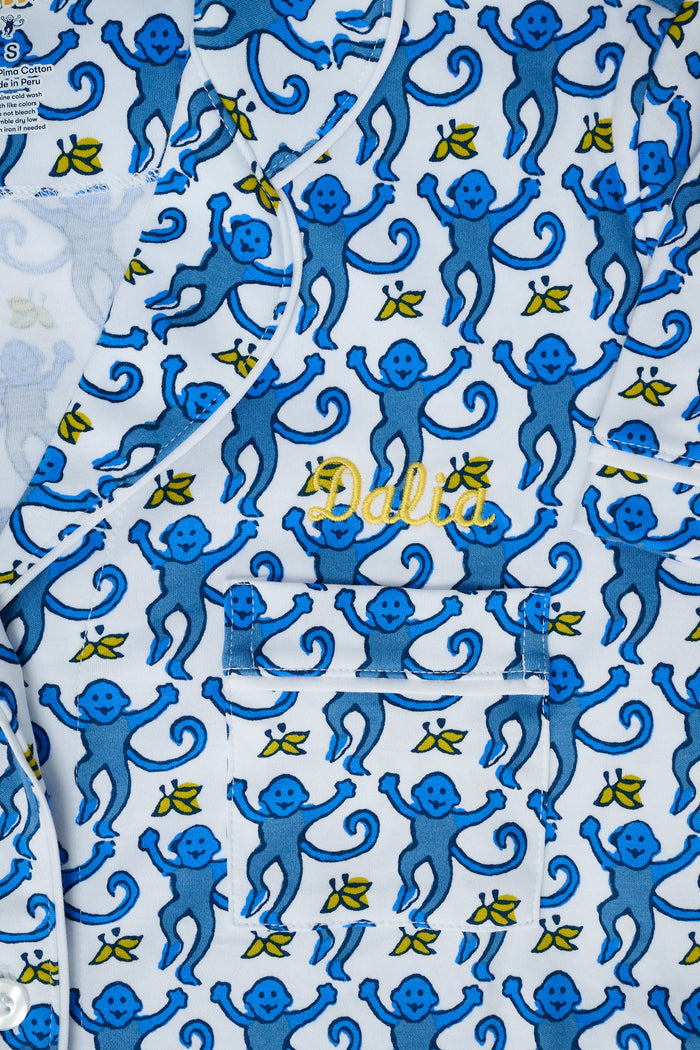 A woman stands barefoot against a plain white background wearing the Roller Rabbit Monkey Fiora Pajamas, a blue-and-white patterned short-sleeve polo pajama set with matching pants.