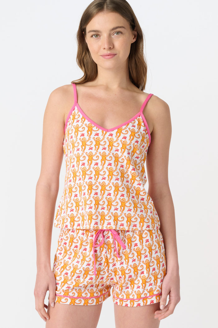 A woman wears the Roller Rabbit Monkey Mischa Set, a white Pima cotton pajama with an orange and pink monkey print, featuring a sleeveless top with pink straps and matching shorts with a pink drawstring, standing against a plain white background.