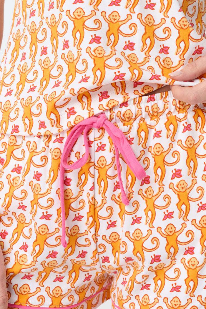 A woman wears the Roller Rabbit Monkey Mischa Set, a white Pima cotton pajama with an orange and pink monkey print, featuring a sleeveless top with pink straps and matching shorts with a pink drawstring, standing against a plain white background.