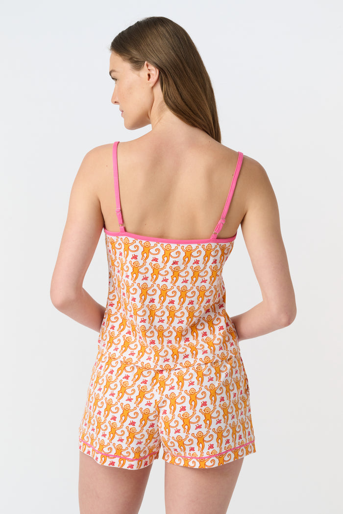 A woman wears the Roller Rabbit Monkey Mischa Set, a white Pima cotton pajama with an orange and pink monkey print, featuring a sleeveless top with pink straps and matching shorts with a pink drawstring, standing against a plain white background.
