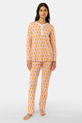 A woman stands barefoot against a plain light gray background in Roller Rabbit’s Monkey Pajamas set, featuring playful orange and white monkey prints, a matching top and bottom, and a white buttoned collar. Her long dark hair adds to the relaxed vibe.
