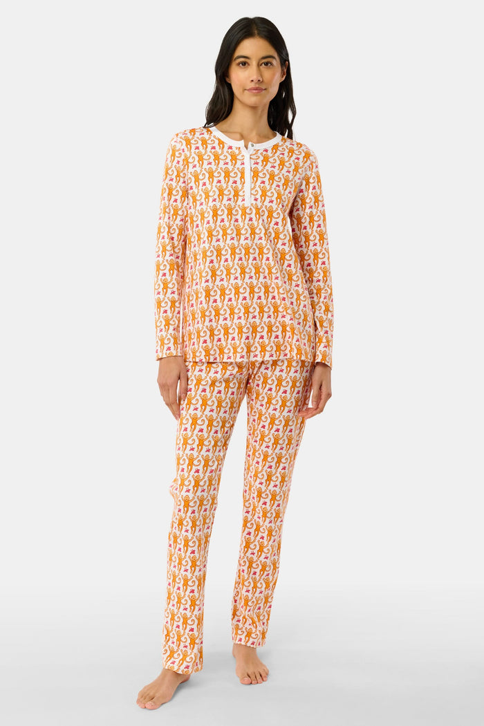 A woman stands barefoot against a plain light gray background in Roller Rabbit’s Monkey Pajamas set, featuring playful orange and white monkey prints, a matching top and bottom, and a white buttoned collar. Her long dark hair adds to the relaxed vibe.