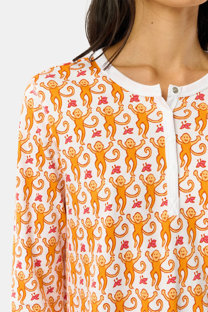 A woman stands barefoot against a plain light gray background in Roller Rabbit’s Monkey Pajamas set, featuring playful orange and white monkey prints, a matching top and bottom, and a white buttoned collar. Her long dark hair adds to the relaxed vibe.