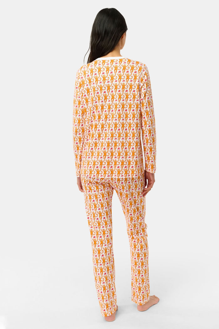 A woman stands barefoot against a plain light gray background in Roller Rabbit’s Monkey Pajamas set, featuring playful orange and white monkey prints, a matching top and bottom, and a white buttoned collar. Her long dark hair adds to the relaxed vibe.