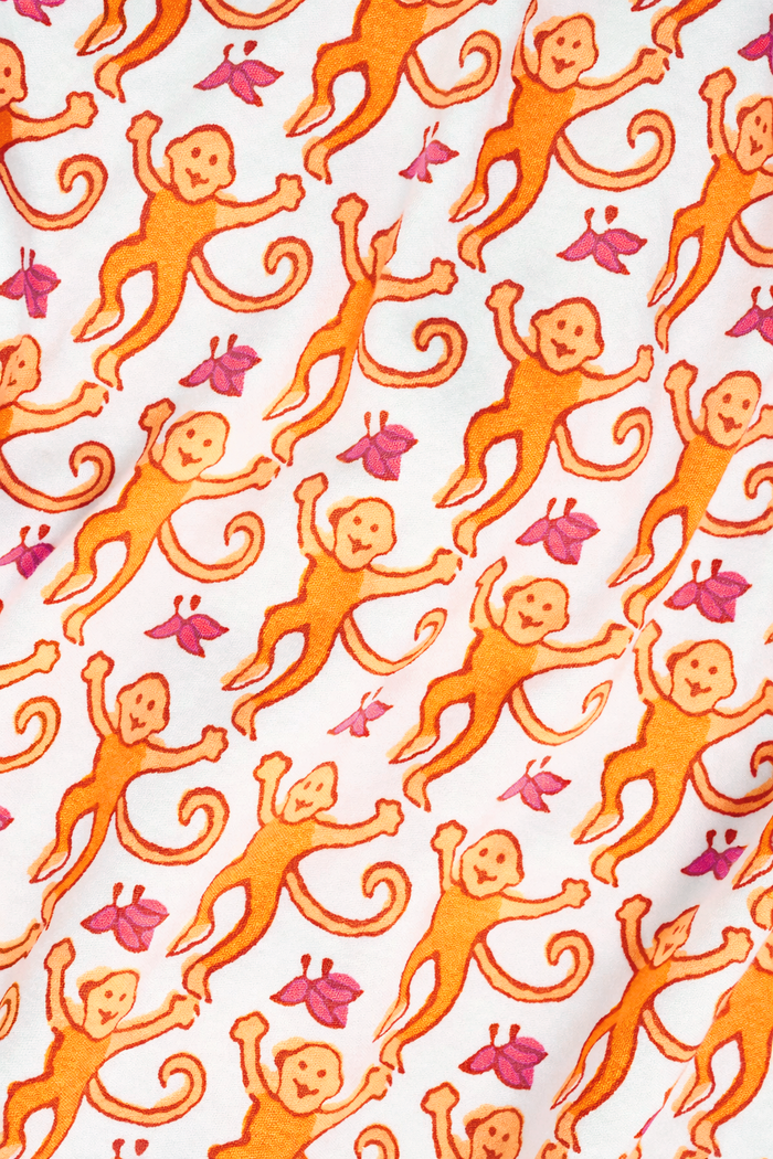 A woman stands barefoot, smiling and looking off to the side, wearing Roller Rabbit Kids Monkey Pajamas—white and orange long-sleeve henley set with playful monkey print and white cuffs.